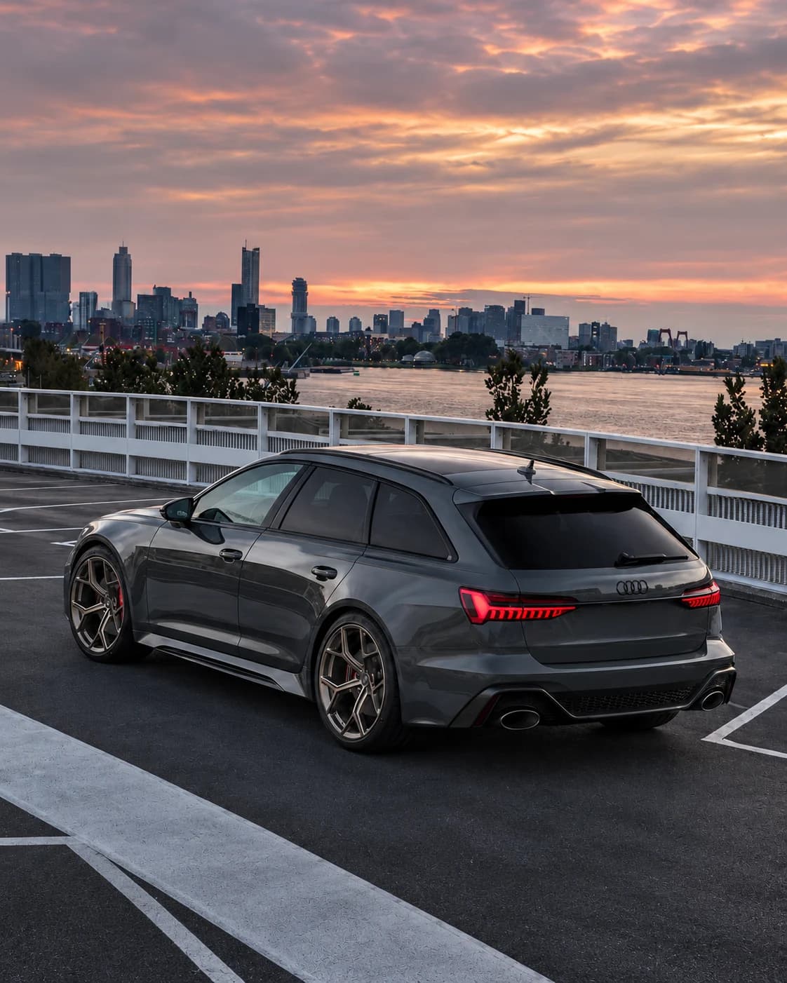 Audi RS6 grise au bord de l’eau, devant une ville au coucher du soleil